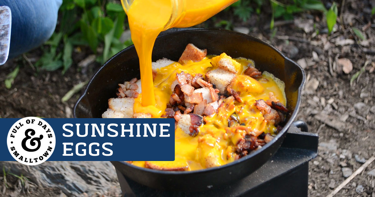 Sunshine Eggs Camping Breakfast, image shows eggs being poured into a cast iron pan with bacon and bread cubes.